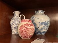 Photo of the three pottery items on a wooden shelf showing red and white ginger jar, blue and white vase, and tall blue and white jug