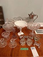 Wide view of lot showing various crystal and milk glass items on wood table including pedestal bowls, lemonade jug, candle sticks, tea-light holders, and covered candy dish.