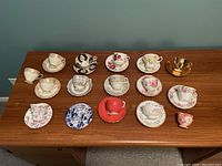 Full view of 13 cups and 11 assorted saucers arranged on wood table showing varied floral patterns, colors, and styles including one gold-tone cup without a saucer.