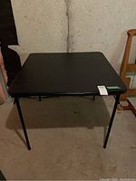 Black square folding card table with padded top, showing the table fully set up with legs extended and square top visible.