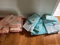 Full view of both coral and teal guest towel sets stacked side by side on floor showing variety of towel sizes and lace/rose embellishments.