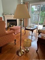 Floor lamp and table lamp side by side on wood floor in living room. Floor lamp with tall cream lampshade and brass base; smaller table lamp also with brass base and cream pleated shade.