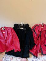 Three coats side by side: red leather jacket with lapel collar, black suede coat with fur collar and matching hat, and red leather and black trim long coat.