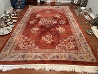 Full view of reddish-brown wool area rug with floral patterns and detailed border, laid out on wood floor, surrounded by various household items.
