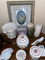 Overview of lot showing framed needlepoint, porcelain jars, decorative dish, candles, and porcelain bird figurine on wooden table