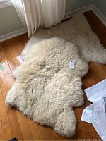 Two natural off-white sheepskin rugs placed on a wood floor under a window, with one rug prominently curly and the other more fluffy, showing texture and natural shape.