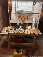 Wide shot of wooden workbench with a variety of hand tools, power tools, clamps, and saws laid out and hanging on pegboard behind, including saws, levels, mitre boxes, clamps, box cutters, masks, Black & Decker electric screwdriver, staple tool, wax polisher, extension cord, and hardware caddy.