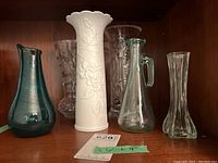 Five assorted decorative vases on wooden surface with auction lot tag visible. Includes white Kaiser vase, green hand blown glass vase, and three clear glass vases.