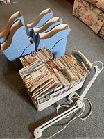 Full view of sewing patterns in a floral cardboard box, four blue magazine organizers behind, and white desk magnifying lamp on floor