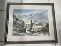 Framed watercolour painting showing a mountain village scene with houses, trees and hills in background, signed by artist.