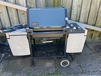 Front view of the Weber gas BBQ grill showing hinged side shelves, attached cooking utensils, and the cooking area under the lid.