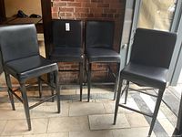 Four black vinyl covered bar stools with metal frames shown against brick wall and tiled floor.