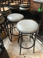 Two metal frame bar stools with round light gray vinyl seats side by side on a tiled floor, showing front and side angles.