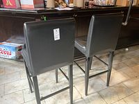 Back view of two black vinyl bar stools showing the square metal legs and the backs of the stools, set against a bar counter and tiled floor.