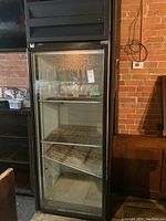 Front view of the upright fridge with glass door, two interior shelves, black exterior against brick wall.
