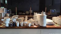 Wide photo showing a variety of stacked cups, saucers, small plates, and bowls, mostly white ceramic with some clear glasses.