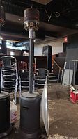 Full view of tall, grey propane patio heater standing upright indoors with stacked chairs in background.
