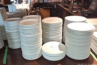 Multiple stacks of white ceramic plates including large dinner plates with wide lips, smaller luncheon plates, and dinner plates with thinner lips.