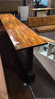 Long wooden table top with warm toned wood surface, mounted on black legs connected by a horizontal base beam.