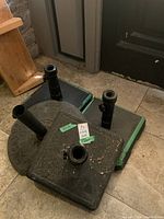 Four black metal umbrella stands with square and round bases stacked together indoors on stone floor near wooden furniture, showing wear and dirt.