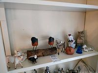 Shelf displaying various animal figurines made of wood, glass, and ceramics including two mounted wooden birds, a white cow, a bald eagle, a white dog with black spots, an owl, a blue glass figurine, and other small ceramic animals.