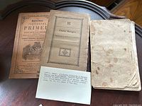 Three antique books laid out on a wooden surface. Titles visible include 'National Pictorial Primer', 'Curtis' Recipes Medical & Miscellaneous', and a heavily stained third book.