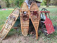 Three wooden snowshoes and one red wooden sled arranged on the ground and picnic table. Snowshoes show woven lattice patterns and leather bindings; sled displays chipped red paint and metal runners.