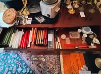 Open drawer showing assortment of candles in multiple colors and sizes, Lexington cutlery sets, and small decorative glass items alongside a paperweight.