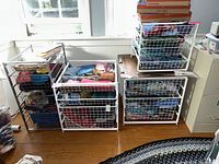 Photo showing four Elfa metal racks with wire baskets holding fabrics, and one black steel 4-drawer metal rack against windowed wall