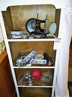 Shelf unit containing a miniature antique child's sewing machine, porcelain trinket boxes, decorative bird-themed trays, glass serving dishes, a red bowl, and a vintage lighter.