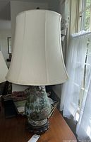 Full view of the Asian hand painted enameled brass base table lamp with cream fabric shade on wooden base.