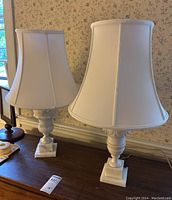 Pair of white marble base lamps with matching white fabric shades sitting on a wooden surface against floral wallpaper.