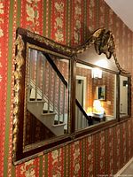 Full view of antique wooden framed mirror with gold leaf decorations and eagle carving at top center, showing wallpaper and adjacent space reflection.