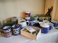 Photo showing assortment of novelty and international mugs in various colors and designs, ceramic trolley sugar bowl, condiment server, and box of cupcake decorations.