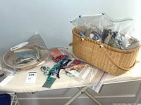 Photo showing needlework hoops, threads, and yarns displayed on an ironing board, including a wicker basket holding some items.