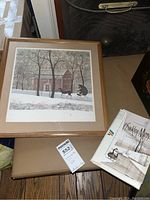 Framed print with winter scene showing silhouette of man on a bicycle and dog, red brick building, trees, and stone wall; P. Buckley Moss book placed beside it.