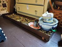 Long metal tray with loose garden stones and items inside including ceramic planters, plate, and watering can.