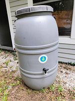 Side view of large gray plastic rain barrel with logo sticker, spigot, and calling out outdoor use condition.