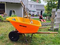 Side view of True Temper bright orange wheelbarrow with wooden handles and single black wheel, set outdoors on grass.