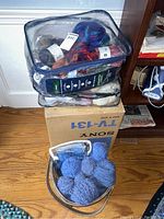 Plastic bag filled with various full and partial wool yarn skeins, some multicolored, placed on a box on a hardwood floor.