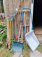 Photo showing full set of yard and garden tools leaning against a wooden barn door outdoors, including rakes, shovels, sledgehammers, pole diggers, and a push plow.
