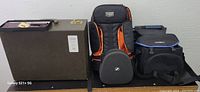 Image showing three assorted camera cases arranged side by side: a large rigid case with metal latches, a black and orange padded Sennheiser case, and a soft compact black fabric camera bag with blue trim.