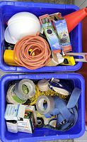 Two blue bins packed with various hardware and workwear items including a white hardhat, orange extension cords, orange traffic cone, levels, tape, nails, ropes, and straps.