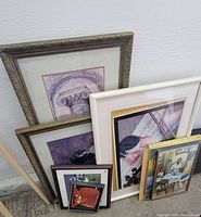 Five framed prints leaning against a wall showing various art styles, colors, and frame designs.