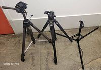 Three extended tripods of different sizes and styles shown standing on floor. Left has metal tripod with large fluid head and handle, middle is carbon fiber tripod with fluid head, right is smaller lightweight tripod stand.
