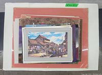Stack of assorted unframed prints on various backing boards with a colorful street scene print on top, size marked 25"W x 18"L