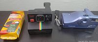 Photo showing Kodak Funsaver disposable camera in unopened packaging, Polaroid Time-Zero Onestep camera with strap, and Polaroid Joycam camera side by side on black surface