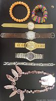 Three wristwatches laid vertically with various bracelets and costume jewelry below on black background.
