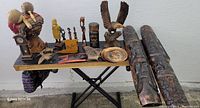 Full display of assorted wooden carvings and two large wooden carved drums on folding table against white wall.