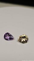 Photo showing two pear-shaped faceted gemstones: golden citrine and purple amethyst on a white background.